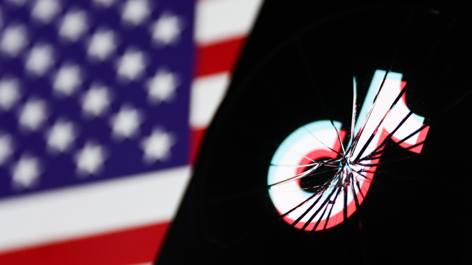 The TikTok logo on a smashed mobile phone screen, behind it the American flag is visible.