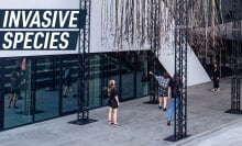 People standing underneath the 'Invasive species' installation which hangs on a metal construction outside a modern building.