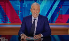 A man in a suit sits behind a talk show desk, grimacing.