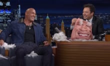 Two men on a talk show are covered in feathers and laughing. One holds a broken cushion.