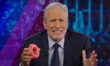 A man in a suit sits behind a talk show desk holding a donut.