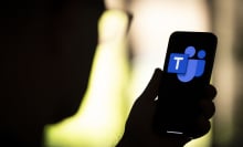 A person in silhouette holds up a phone displaying the blue Microsoft Teams logo.