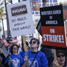 A group of people wearing blue shirts walk down a Hollywood street carrying picket signs. One sign reads "Writers Guild of America on strike!" Another sign reads "WGAW. No pages without fair wages."