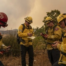 Four firefighters stand in a circle looking down at radios and notepads. 