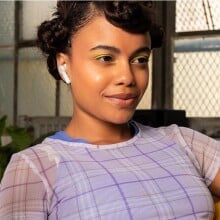 a person with earbuds in gives a slight smile while looking at a computer screen. She also has a phone with a yellow case in her right hand.