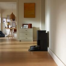 Black Roborock Qrevo S robot vacuum on self-empty dock sitting against wall with living room scene in background