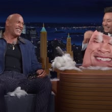 Two men on a talk show are covered in feathers and laughing. One holds a broken cushion.