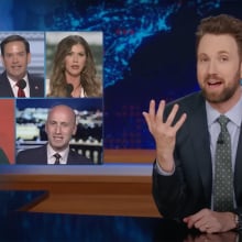 A man in a suit sits behind a talk show desk, gesturing at images of five other people in the top-left.