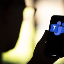 A person in silhouette holds up a phone displaying the blue Microsoft Teams logo.