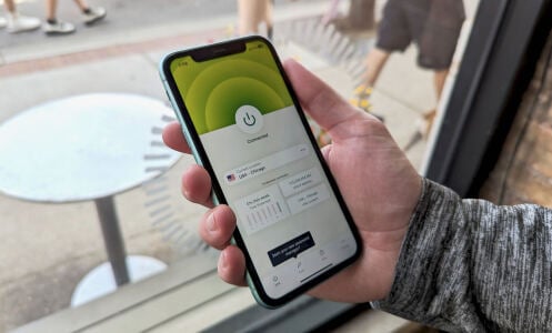 a close-up of a man holding an iphone displaying the expressvpn app in front of a window