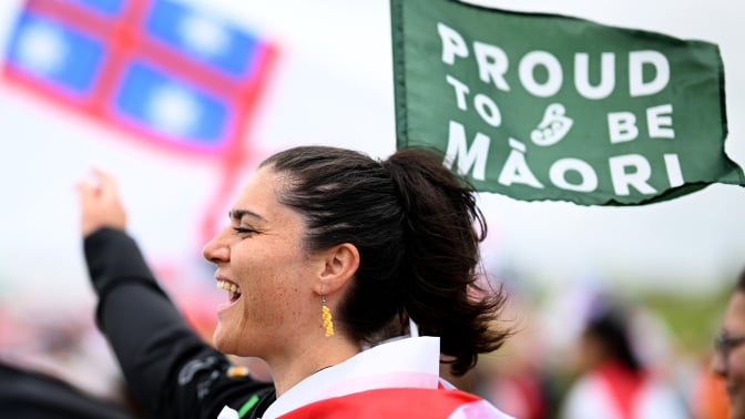 A person cheers and holds up a green flag that reads "Proud to be Maori."