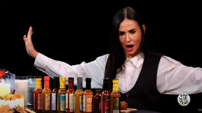 A woman throws her arms wide while sitting in front of a table covered in sauce bottles.