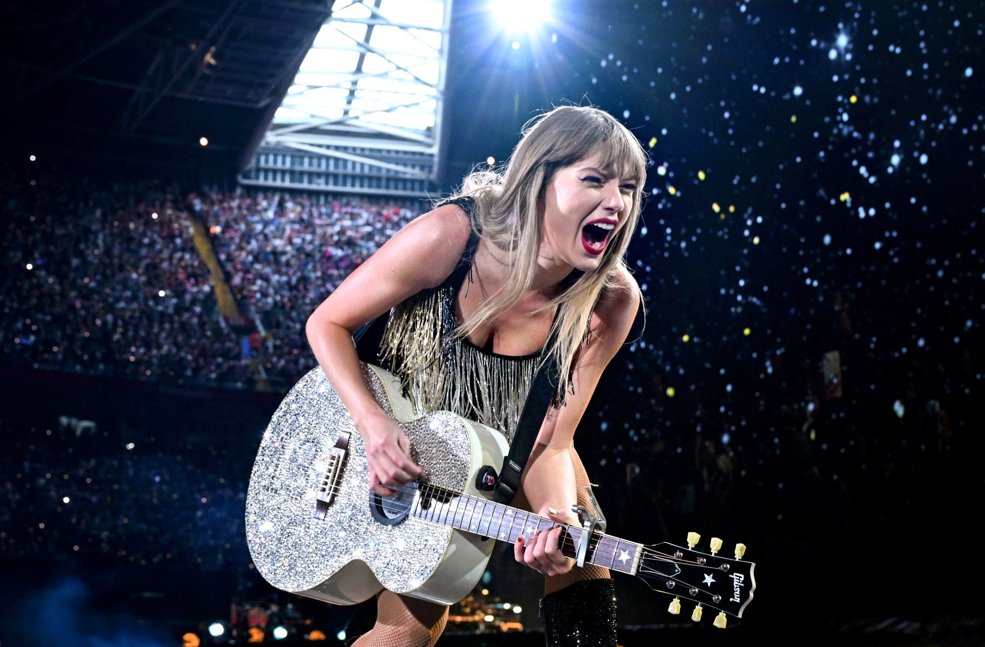 Taylor Swift playing a rhinestoned guitar, mid-concert.