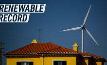 A picture shows the top of a typical portuguese house, yellow walls, dark green shutters, and a red rooftop. Behind it, against the blue sky a win turbine is peeking in the background.