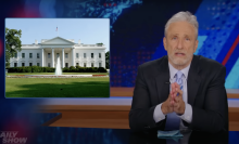 A man in a suit sits behind a talk show desk with an image of the White House on the left.