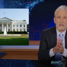 A man in a suit sits behind a talk show desk with an image of the White House on the left.