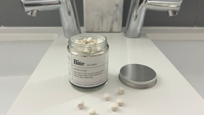 Bite Toothpaste jar with lid off and Bits on countertop with faucets and toothbrushes in background