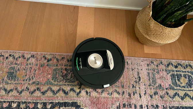 Shark robot vacuum cleaning rug with hardwood floor, wall, and plant in peripheral