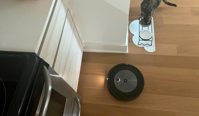 iRobot Roomba Combo j5 robot vacuum cleaning hardwood floor with kitchen counter, stove, and cat eating food in peripherals